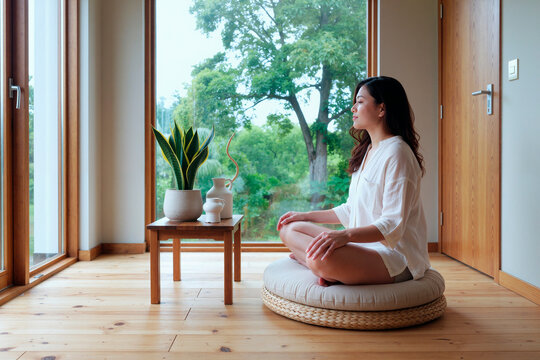 Asian young woman meditating cross legged by window, practicing mindfulness and stress relief at home. Ideal for mental wellness, self care, meditation app or therapy marketing