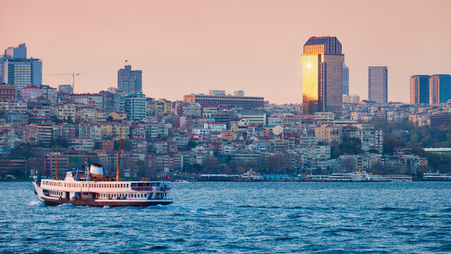 Panoramic sunset view of Istanbul, Turkey, with modern towers and traditional architecture along Bosphorus. Ferries and boats are visible on water in foreground during sunset