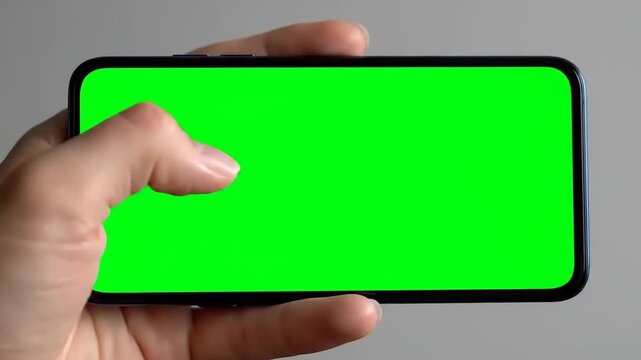 Close-up view of a hand holding a modern smartphone horizontally, displaying a vibrant green screen for easy content overlay and digital compositing in post-production