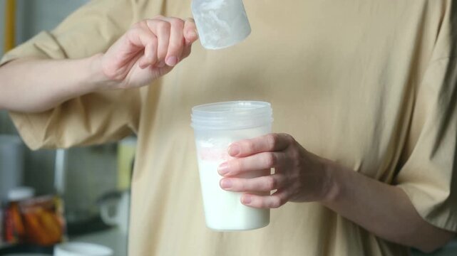 Female hands pouring scoop of white protein powder supplement into shaker bottle with milk or water to prepare post workout nutritional drink for muscle gain and recovery.