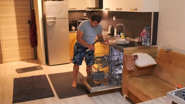 Man removing kitchen utensils from sliding dishwasher rack after washing cycle, organizing clean cutlery and tools, everyday household routine in modern apartment kitchen interior