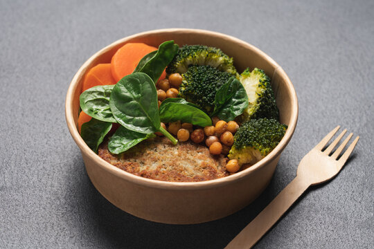 Healthy vegan lunch with broccoli, roasted chickpeas, spinach, carrot and lentil patty in round cardboard container on black background with copy space