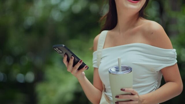 Close-up of a smiling young Asian woman using a smartphone and holding a reusable tumbler in a green outdoor park. Modern eco-friendly lifestyle, mobile connectivity, and sustainable daily
