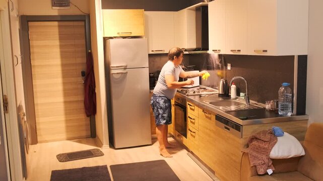 middle aged white man wiping backsplash surface medium view in kitchen, cleaning routine using cloth and detergent, removing grease and stains, hygiene focused domestic maintenance task