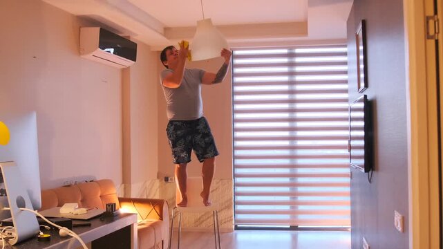 middle aged white man wiping ceiling lamp low angle in living room, cleaning routine using cloth, removing dust and dirt, careful maintenance of light fixture with everyday domestic work at home