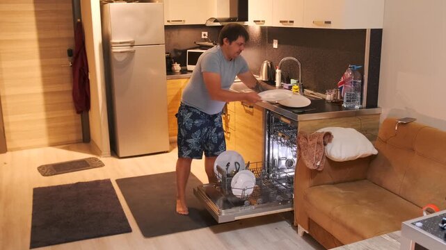Man removing clean plates from dishwasher after washing cycle, unloading kitchen appliance and organizing dishes, everyday household routine in modern apartment kitchen interior
