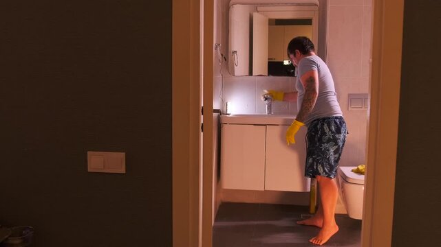 Man scrubbing bathroom faucet with foamy sponge, removing limescale and dirt from metal surface near sink, maintaining hygiene and cleanliness in modern home bathroom interior routine (