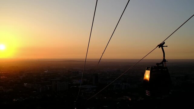 Cable car gondola over Almaty city skyline at dramatic orange red sunset dusk