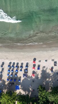 Imagem a&eacute;rea da orla de praia com ondas do mar, areia e guarda-s&oacute;is organizados, representando turismo e lazer em dia de ver&atilde;o