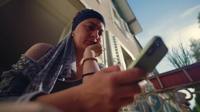 Woman undergoing chemotherapy low angle close up using smartphone on balcony expressing focus, contemplation, concern and digital connection during challenging health situation