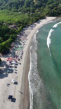 Imagem a&eacute;rea da Praia da Barra do Sahy com faixa de areia ocupada, mar calmo e vegeta&ccedil;&atilde;o ao redor, representando destino tur&iacute;stico popular no litoral paulista