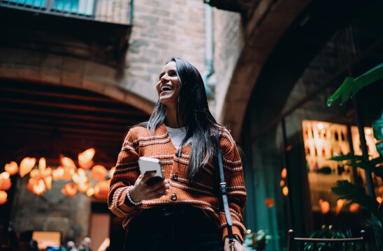 Laughing hispanic woman holding smartphone in city courtyard, happy multiracial girl looking up with joy, young female using mobile device outdoors, authentic expression of happiness and luck.