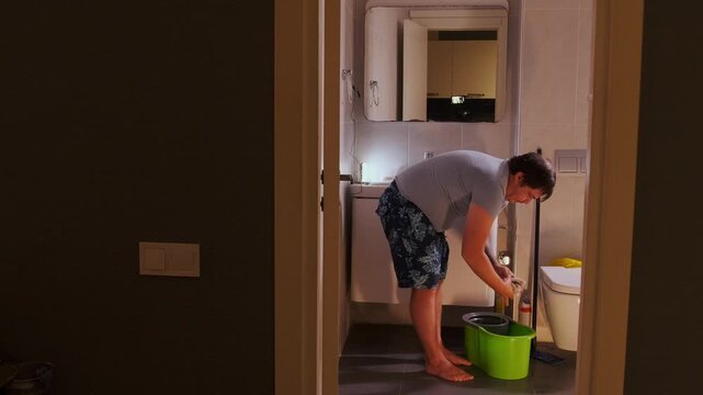 middle aged white man wringing cloth over bucket low angle in bathroom, home cleaning routine with manual effort, household chores scene focused on hygiene, maintenance and everyday domestic work
