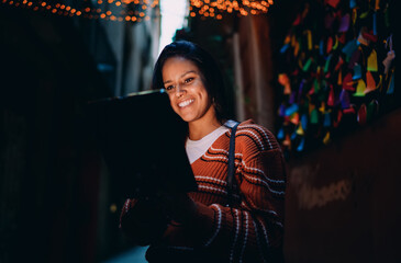 Radiant mixed race woman with tablet at night, city lights bokeh, smiling face, successful online business, financial freedom, digital communication, mobile app usage, authentic lifestyle portrait. © BullRun
