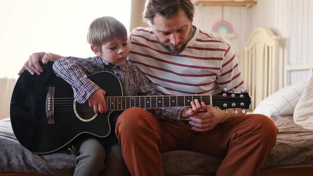 Handsome dad teaches his cute son to play the guitar sitting on sofa in the living room at home. Happy Father's Day. Father teaching his child to play guitar. Kid Learning music skill. Art education