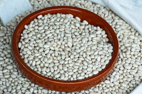 Dry white navy beans filling a brown ceramic bowl on a rustic market display. High-quality top view showing texture and abundance for healthy eating and agriculture themes