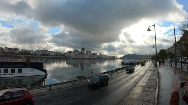 A picturesque river scene featuring a blend of urban architecture, moving vehicles, and an expansive sky filled with clouds. The tranquil water reflects the surrounding buildings, creating a serene ye