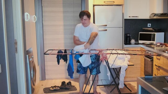 Man shaking and hanging wet clothes on metal drying rack in apartment kitchen, home laundry routine, spreading garments for air drying, everyday domestic chore, indoor lifestyle activity
