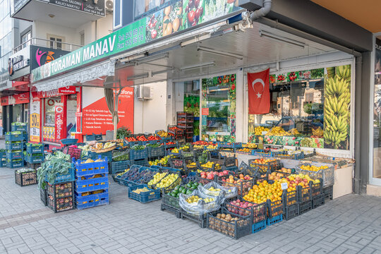 A horizontal eye-level shot features Toraman Manav, a vibrant outdoor fruit and vegetable market. Fresh produce sits in colorful crates beneath a Turkish flag. Alanya, Turkey - October 25, 2020