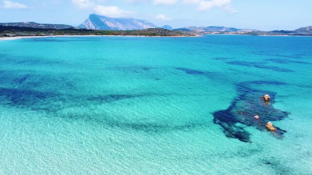 Aerial orbit around rocky formations at Lu Impostu beach in Sardinia, revealing turquoise shallow waters, white sand and coastal textures in clear Mediterranean light.