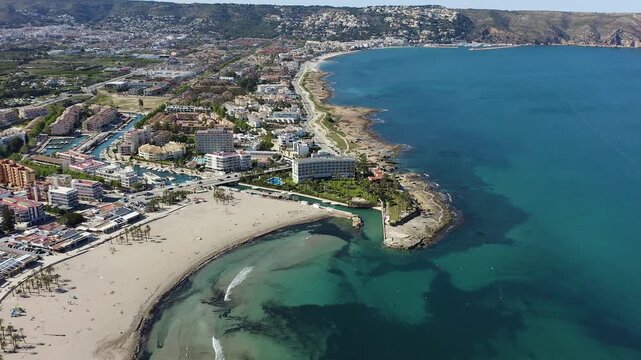 Aerial view of semicircular Arenal beach in J&aacute;vea, Alicante, Spain. Sandy shoreline with calm waters and entrance channel to marina, surrounded by urban coastal landscape. 