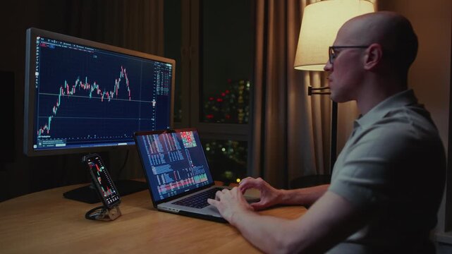 Man trader monitors market charts on laptop, screen and smartphone at home. Analyzing real-time trading data, considering investment strategies. Professional investor engaged in day trading, Evening