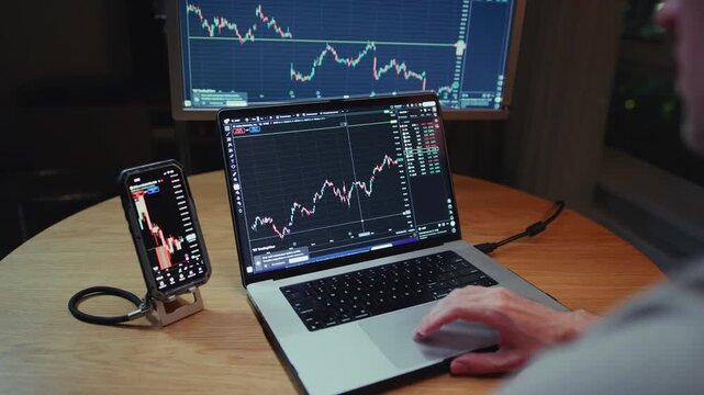 Man trader monitors market charts on laptop, screen and smartphone at home. Analyzing real-time trading data, considering investment strategies. Professional investor engaged in day trading, Evening