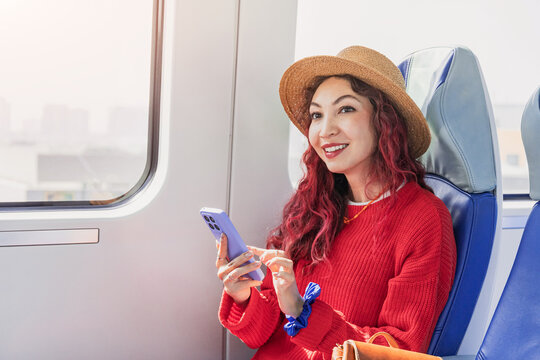 Smiling woman with red hair wearing a hat, sitting by a window on a train, and looking away while holding a smartphone