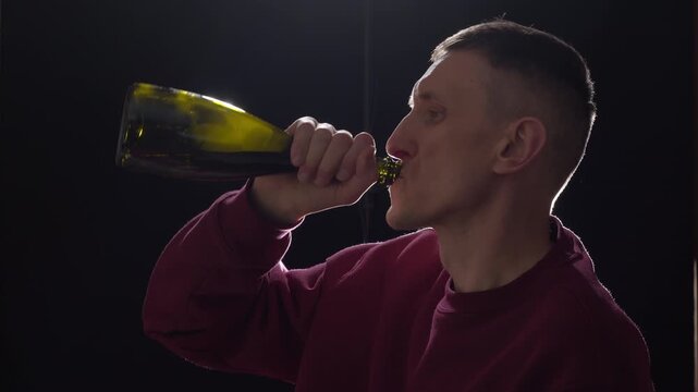 A man in a dark room drinks wine from a bottle. Concept of alcoholism and drug addiction.