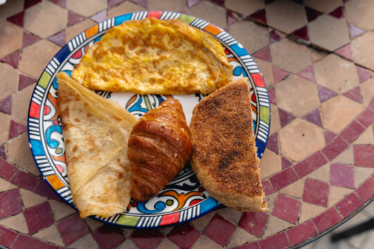 Traditional Moroccan breakfast serving msemen flatbread, harcha, omelet, and croissant on a colorful mosaic table