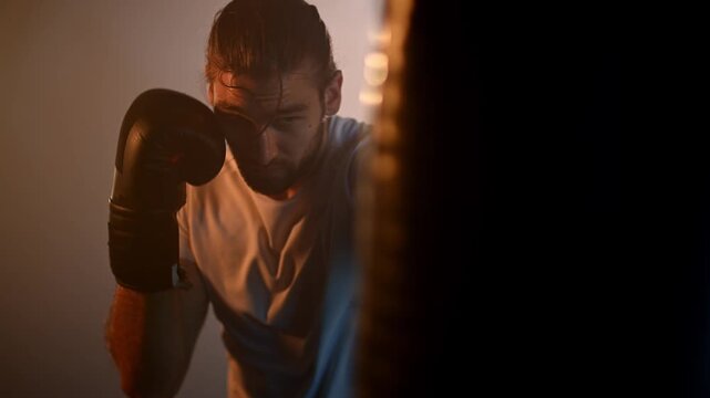Strong sweaty fighter working out with a punching bag in a dark gym during a boxing practice with atmospheric cinematic light