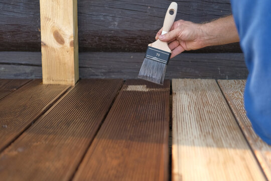 Brown oil for protection of wood by staining. Painting a larch for protecting and pests. Woodworker applying finish to terrace deck board. Repairman varnishing with brush wooden plank floor surface.