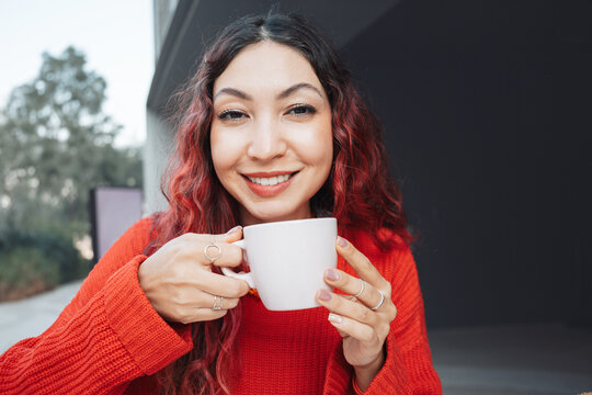 Young woman with red hair and a red sweater, smiling and holding a coffee cup, enjoying a morning drink