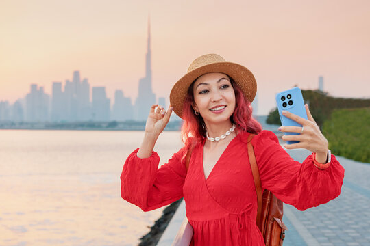 Young woman in red dress and straw hat capturing a selfie in front of the Dubai skyline during sunset