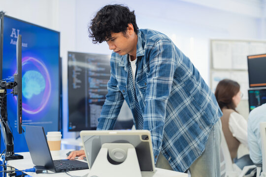 focused young Asian male programmer with curly hair is working intently on his laptop in a modern data center office. The background features bright blue neon LED-lit monitor
