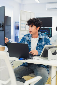 focused young Asian male programmer with curly hair is working intently on his laptop in a modern data center office. The background features bright blue neon LED-lit monitor