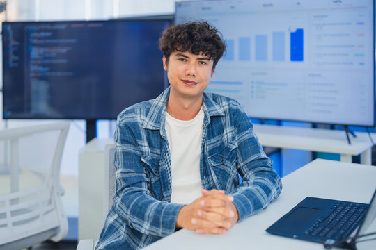 portrait of a confident young Asian male professional sitting at his desk in a modern tech office, looking directly at the camera in a POV style as if participating in an online video conference