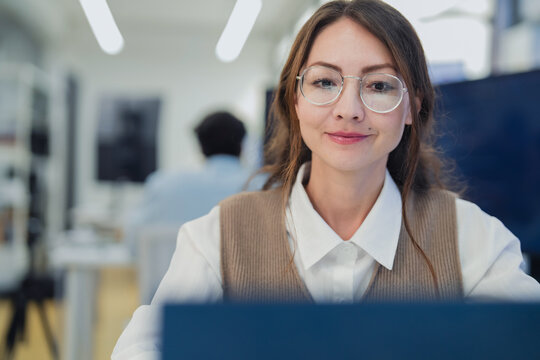 professional female software developer with glasses sitting at her desk, holding a coffee cup while intently working on her laptop. The scene captures a modern business environment 