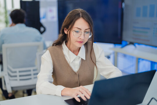 professional female software developer with glasses sitting at her desk, holding a coffee cup while intently working on her laptop. The scene captures a modern business environment 