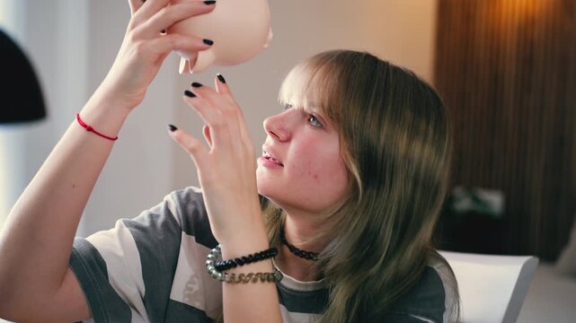 Broke teenage girl trying to get last coins out of piggy bank, symbolizing financial hardship, struggle of saving money, poverty, rising cost of living