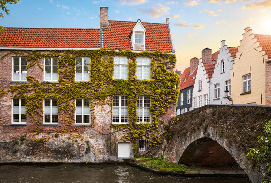 Bruges, Belgium. Medieval vintage brick houses with windows over river with old stone bridge, covered with green plants and bushes. Summer landscape with sky and clouds..