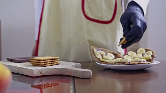 A close-up shot of a person wearing black gloves and an apron, preparing a sweet dessert. They crumble biscuits over a base of banana slices and chocolate spread, creating a delightful treat.