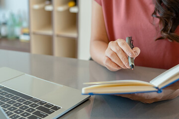 Fototapeta premium Smiling Asian Woman Writing in Notebook by Laptop in Kitchen