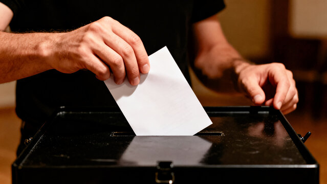 Man's hand inserts a white ballot paper into a black ballot box. His right hand holds the paper, left hand steadies the box. The ballot box features a narrow opening for paper insertion