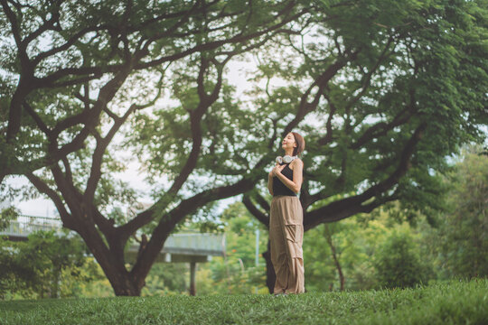 Young Asian woman practicing mindfulness and deep breathing in a lush green park under large trees. Wearing headphones, she enjoys a peaceful moment of mental wellness and nature connection