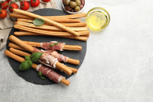 Grissini breadsticks with prosciutto, tomatoes, oil, olives and basil on light table, flat lay. Space for text