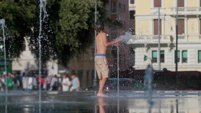 Boy standing barefoot on wet pavement playing with water fountain jet in public square during summer day surrounded by splashes and blurred urban background