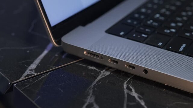 Close-up of hand unplugging magnetic charging cable from laptop port on desk, demonstrating safe quick-release connector, modern device usage, power disconnection, everyday tech interaction