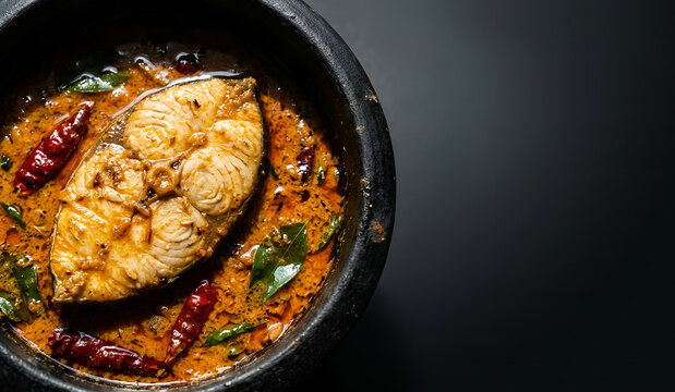 A dramatic, low-key macro shot of one large piece of Kingfish steak in a vibrant, bright orange Goan coconut and tamarind fish curry, served in a black earthen pot on the left 
