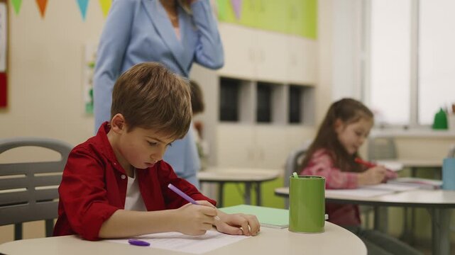Schoolboy writing test in classroom while teacher helping students nearby